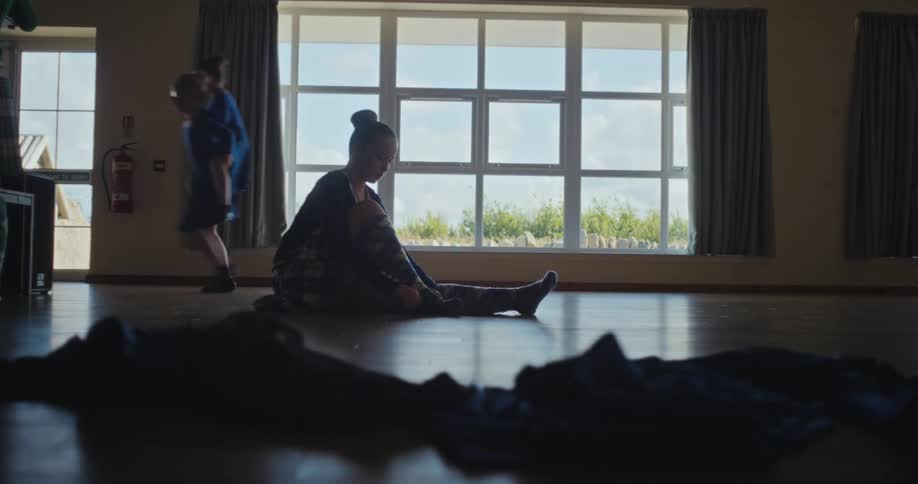 The video shows a group of children in a studio with large windows and a wooden floor in the foreground a girl is stretching while in the background two children are walking with a third child on their back