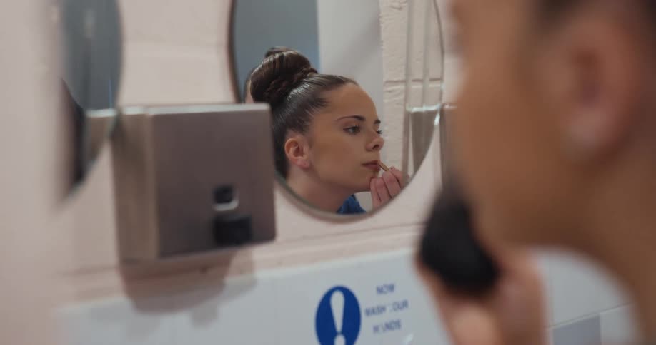 A woman looks into a mirror and applies lip color to their mouth while other woman is doing makeup