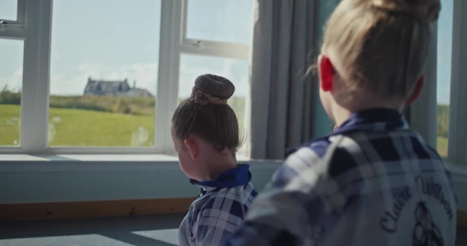 In a sunlit room a young girl with her hair in a bun gets help from another girl who is adjusting her hair with a brush there are other children in the background also with their hair in buns