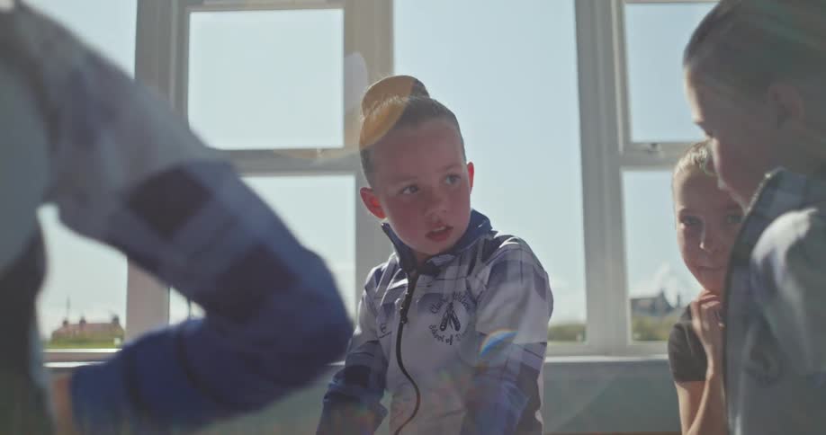 A group of children sitting are talking with each other in a room with a window in the background