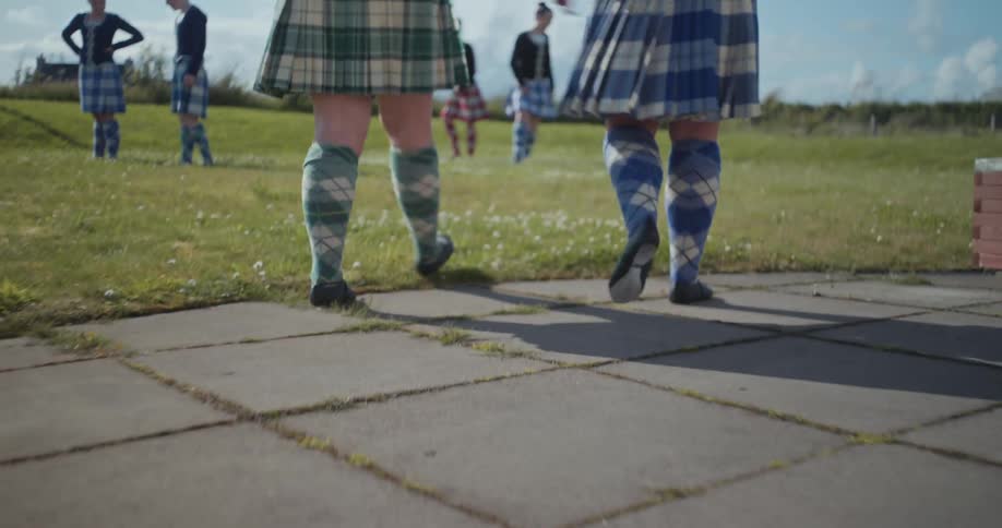 Dancers in kilts rehearse outdoors on a lawn under the sunny sky