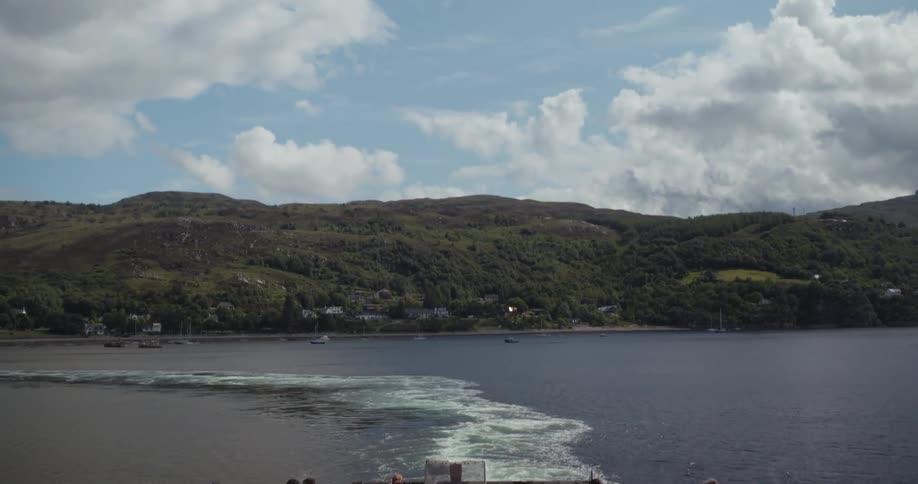 Passengers on a ferrys deck gaze out at a beautiful mountainous coastline with scattered buildings as the boat leaves a white wake behind it