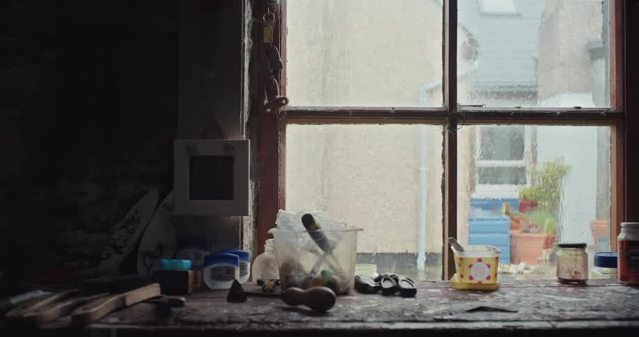 Rain hits the window of a room with a cluttered desk