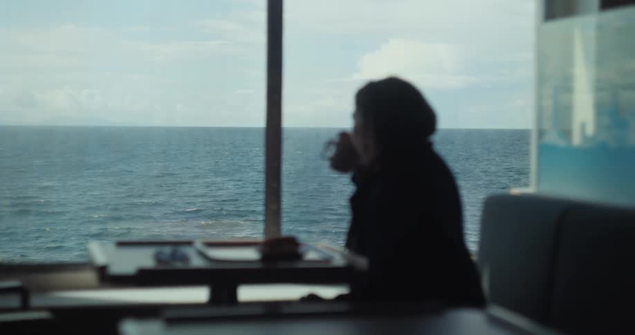 Person drinks a beverage while gazing at the sea through a large window