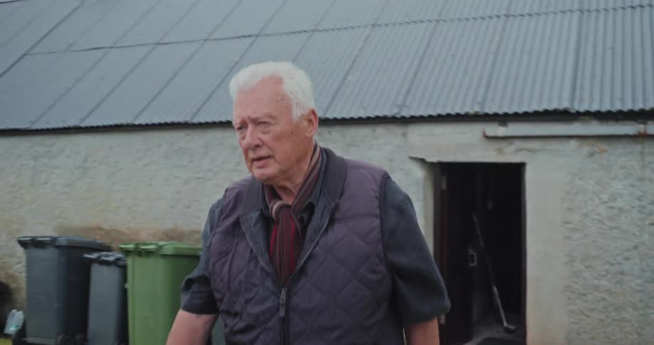 An elderly man with white hair and a gray beard emerges from a dark doorway and walks across a farmyard he wears a dark vest over a shirt and has a scarf around his neck