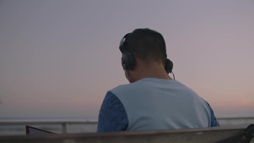 Man wearing headphones sits on a bench in front of water