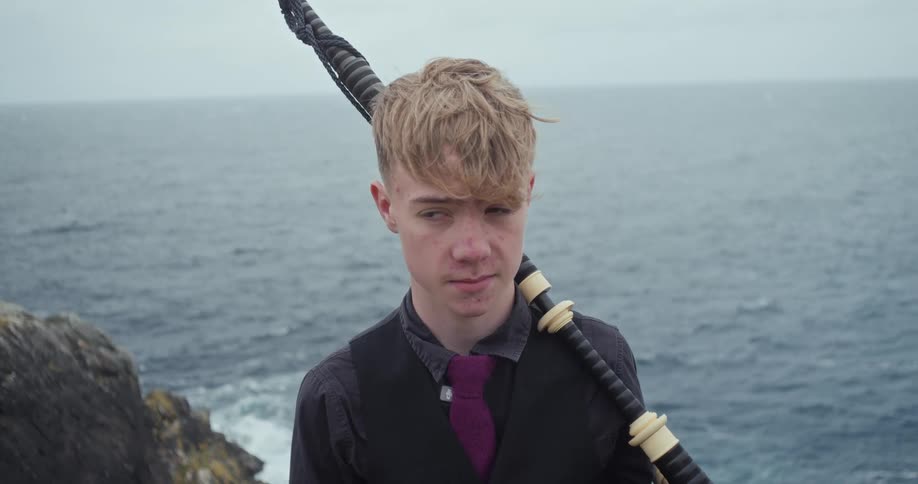 The man is standing near the water with bagpipes instrument