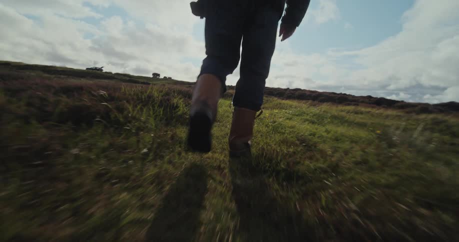 A person wearing a dark jacket and jeans is walking through a grassy field the sun is bright and low in the sky creating a silhouette of the person as they walk away from the camera