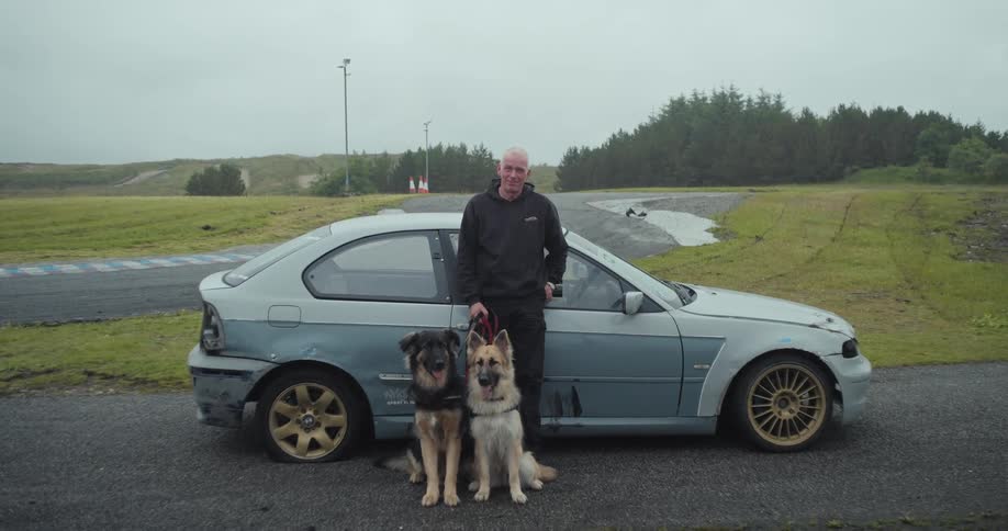 A man stands holding leashes for two dogs with a car parked next to him on a paved surface
