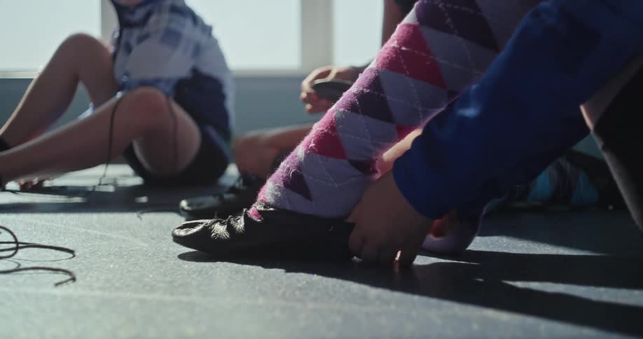 A young person wearing patterned argyle socks is seen sitting on the floor lacing up a pair of black dancing shoes other dancers are in the background also getting ready with one persons hands visible as they adjust their footwear