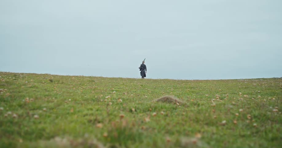 Man walks alone in an open field carrying a tripod on his shoulders