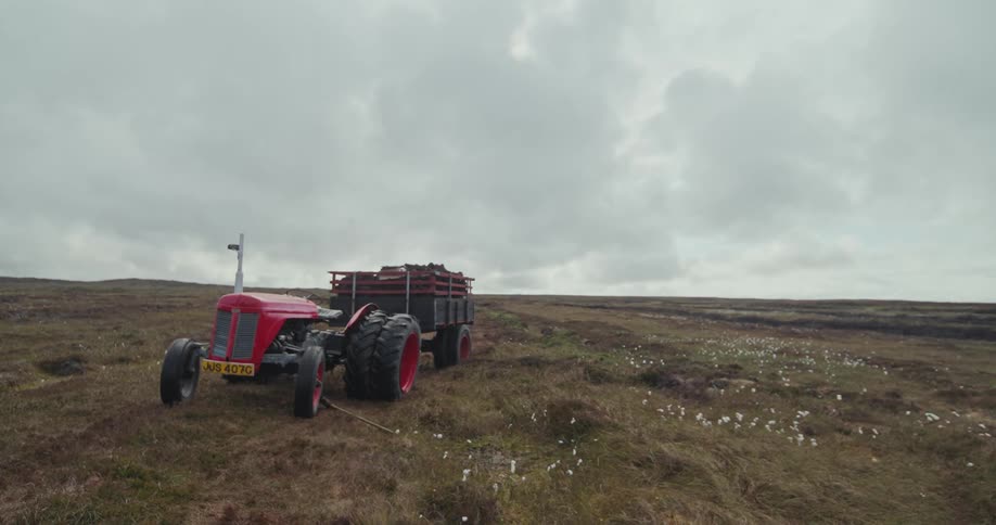 A full view of an old red tractor with a trailer in a field of dry grass under a dark cloudy sky