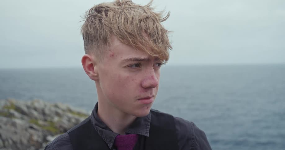 A person with a neutral face looks down and to the side while standing in front of the ocean