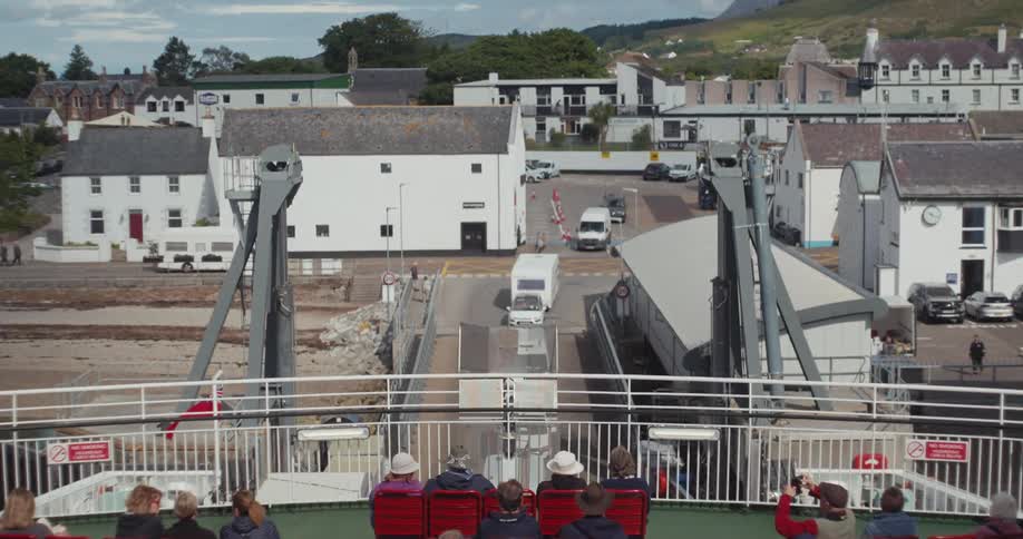 A full view shot from the deck of a ferry shows people sitting in red seats as the ship approaches a small town with white buildings and green hills
