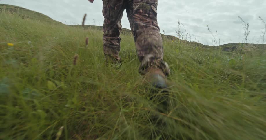A person wearing camouflage pants and black boots is seen walking through tall green grass on a hillside the camera is low to the ground capturing the movement of their legs as they walk