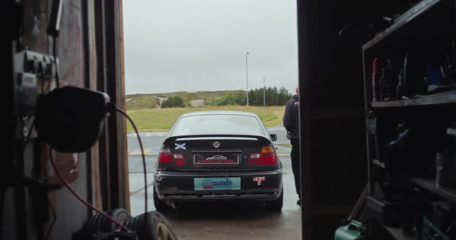 A man in a black hoodie and pants walks from a small shed with a helmet and gets into the drivers seat of his black race car