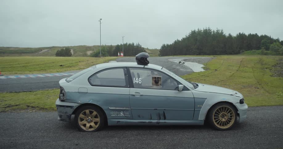 A low angle view shows a modified white and gray race car with a helmet on its roof and a dog in the passenger seat parked on an asphalt track with grass and trees in the background