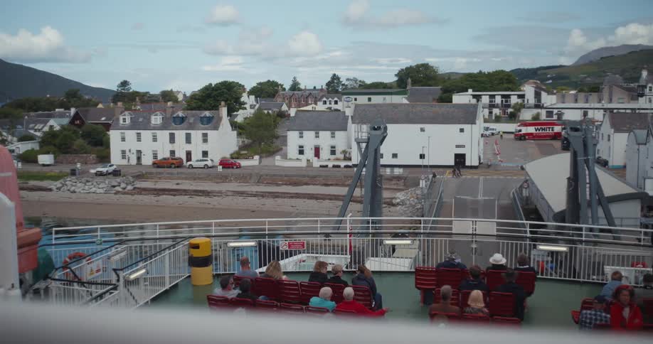 A ferry docks at a port town with passengers seated on deck and white buildings nestled against a mountainous backdrop