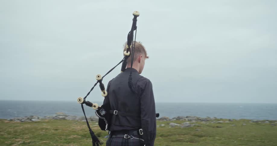 A person with a bagpipe over their shoulder walks across a grassy field toward the ocean