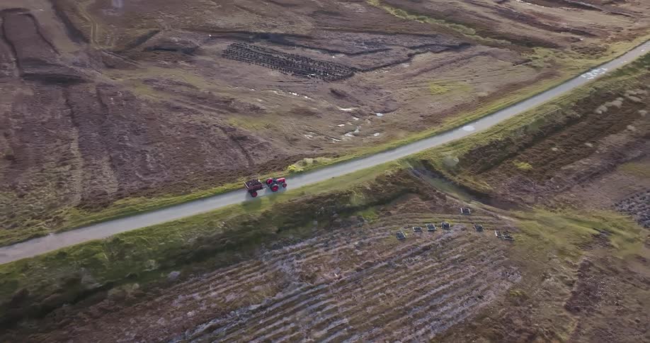 The video shows two tractors driving along a road on what appears to be a moor below the road there are rows of a crop planted in the soil