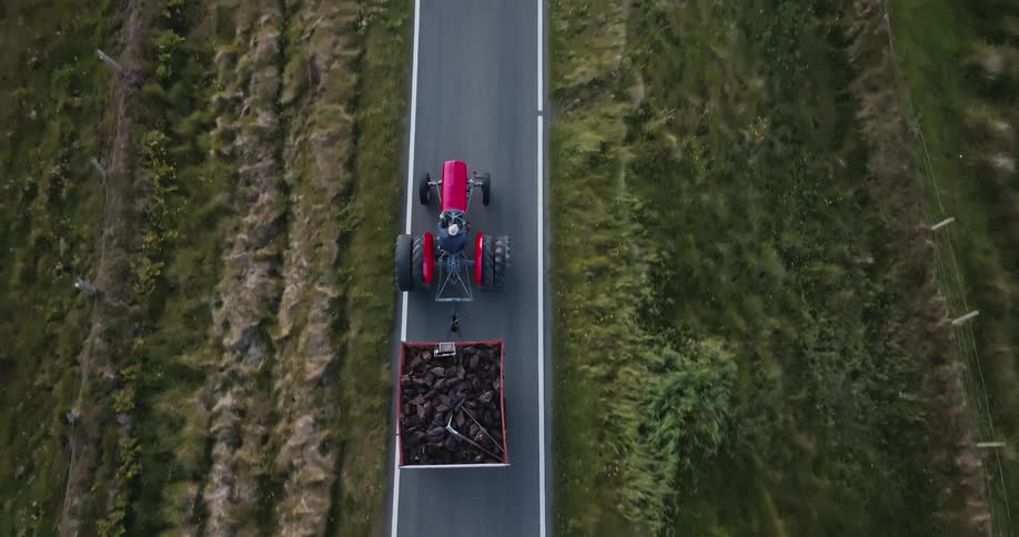 A red tractor pulls a trailer full of peat down a narrow road surrounded by green fields