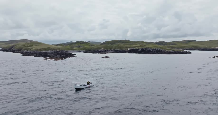 A small white boat with one person travels across a choppy sea toward a rocky green coastline under a cloudy sky