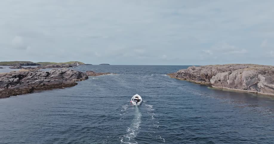 A single small boat travels on dark blue water between two large rocky islands on a cloudy day leaving a white wake behind it