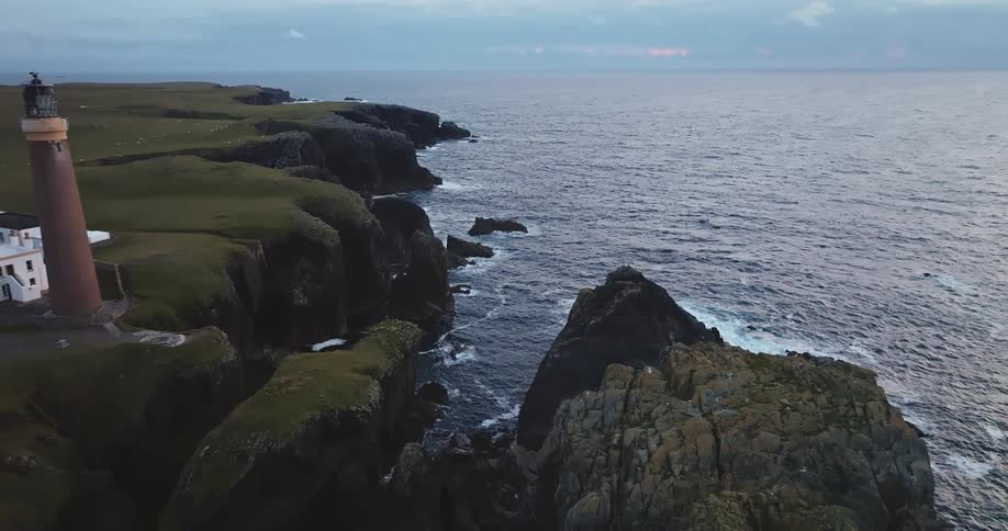 A lighthouse stands on a rugged grassy cliff overlooking a vast dark ocean under a cloudy sky