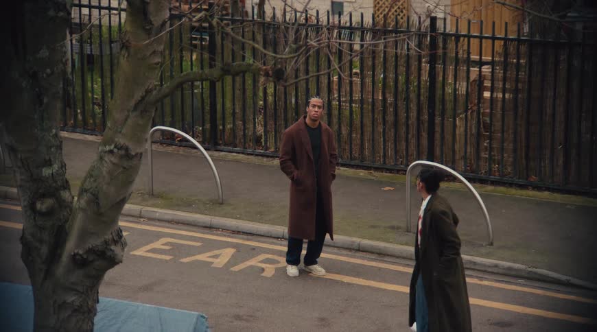 A man and a woman in winter coats are walking on a city sidewalk when the man pauses and the woman convinces him to keep going