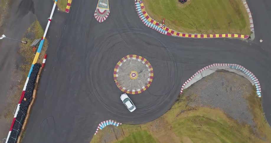 A white sedan performs a continuous burnout spinning in a tight circle on a gray asphalt racetrack with painted white and red curbing