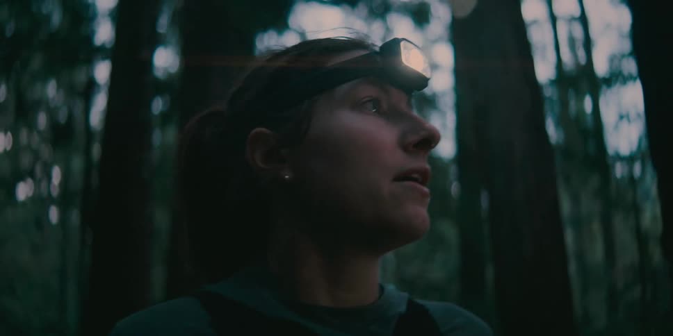 A female hiker turns on her headlamp while standing in a forest at dusk