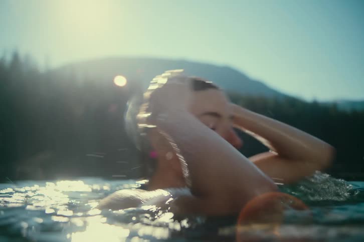 A woman emerges from a body of water with water droplets on her face and hair the sun is setting behind her creating a bright reflection on the waters surface and highlighting the landscape in the background