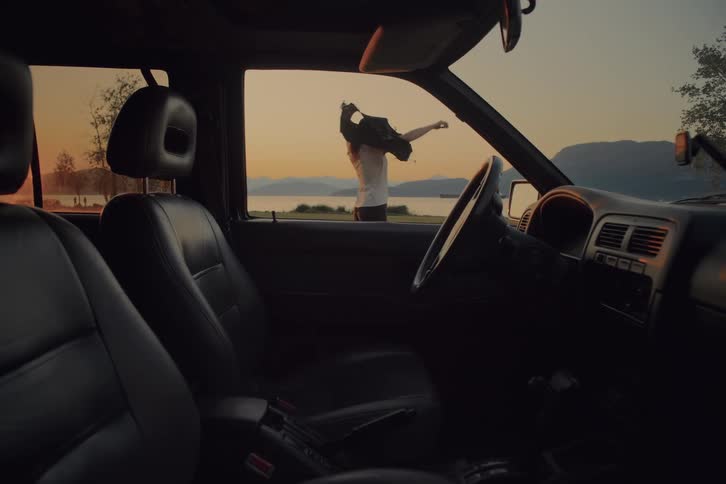 A view from inside a car shows a woman in a white shirt and black shorts with her back to the camera as she walks out the open door and stands near a lake and mountains at sunset
