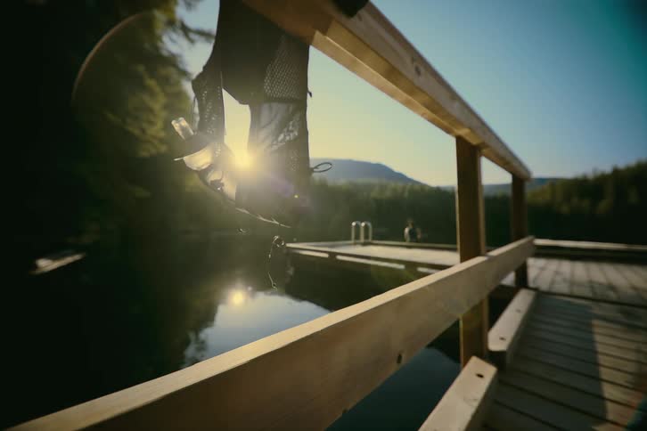 A running vest hangs from a wooden railing on a dock as a woman jumps from the dock into a lake
