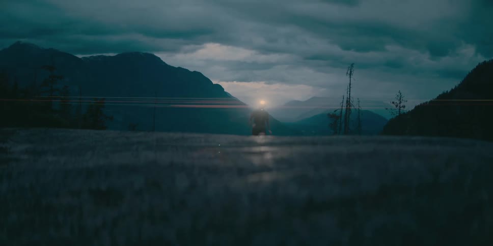 In the clip a person wearing a headlamp runs through a field at dusk the dim light from the headlamp is visible as they jog across a grassy area surrounded by a serene mountainous landscape under a cloudy twilight sky