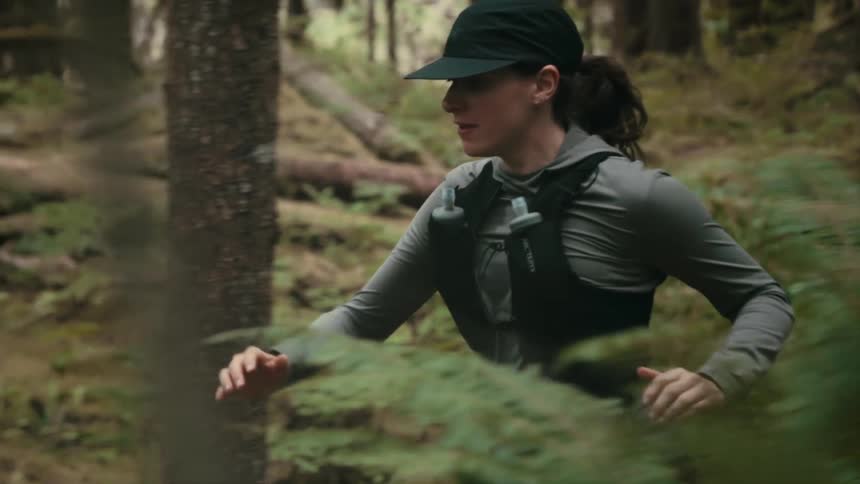 A low angle close view shows a woman wearing a gray long sleeve shirt black running vest and a cap as she jogs past green ferns in a dark forest