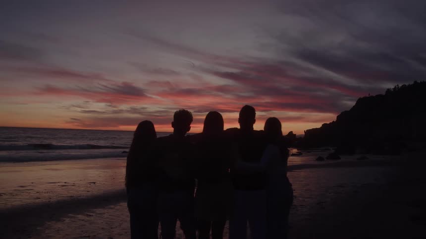 Group of five friends in silhouette hugging each other at the end of the afternoon at the sea