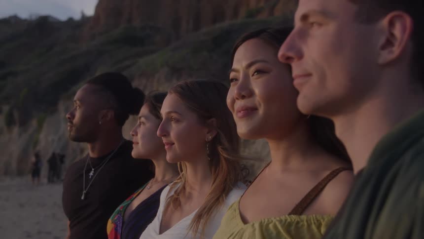 A group of five friends is standing together on a beach at sunset looking out at the water they are sidebyside with the last light of the day illuminating their faces