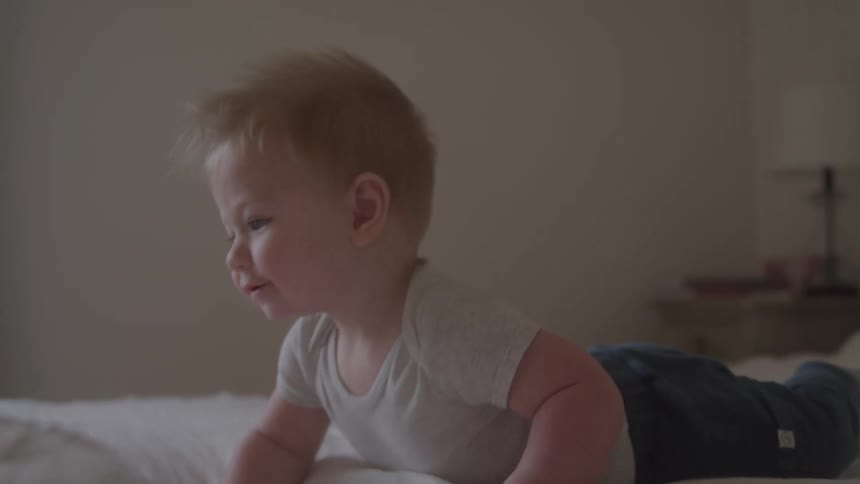 In the first video a person in a white spacesuit walks through a futuristic redlit hallway in the second video a happy smiling baby is seen crawling and playing on a white bed