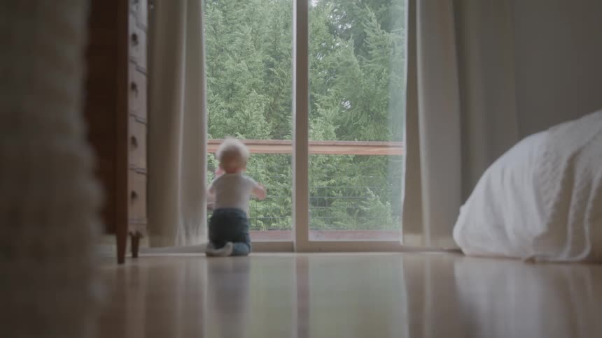 A baby crawls on a wood floor toward a large window and then rises to stand