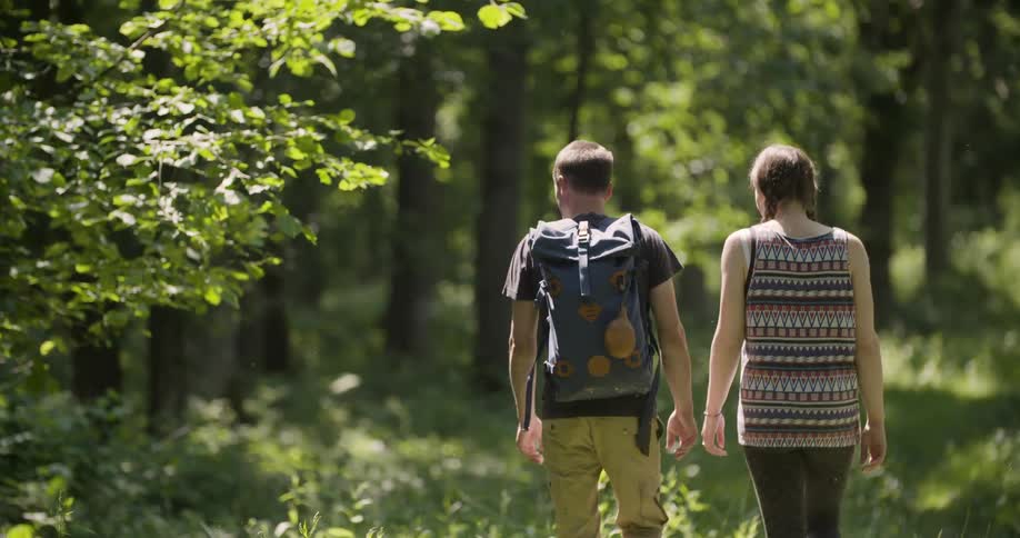 A couple of teenagers walk away from the camera through a sundappled forest with green grass and bright green leaves on the trees