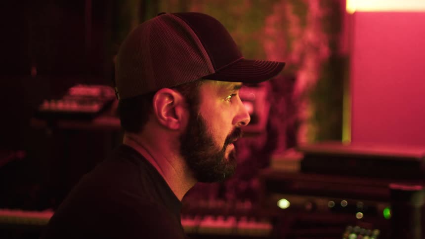 A bearded man in a dark cap sits in a dimly lit room appearing focused and contemplative