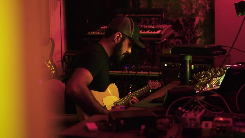 A man plays electric guitar in a dimly lit studio surrounded by equipment
