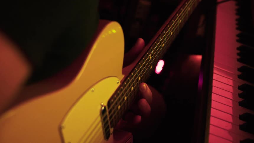 A close view of a persons hands playing chords on a yellow electric guitar with a piano keyboard and a dark redlit background visible