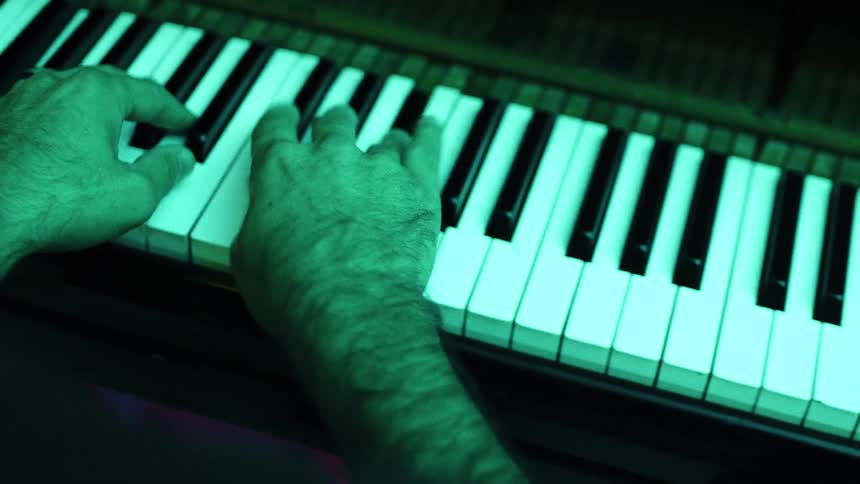 A top view of a mans hands playing chords on a piano with a green light cast over the keyboard