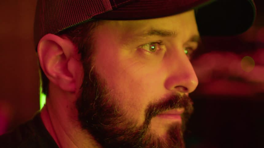 A bearded man in a cap looks focused and serious under warm lighting