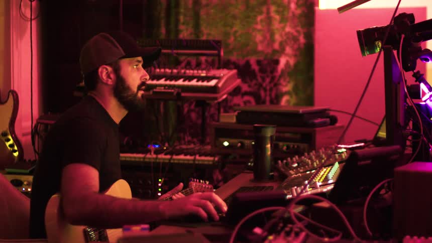 A man with a guitar works at a sound mixing desk in a dimly lit studio