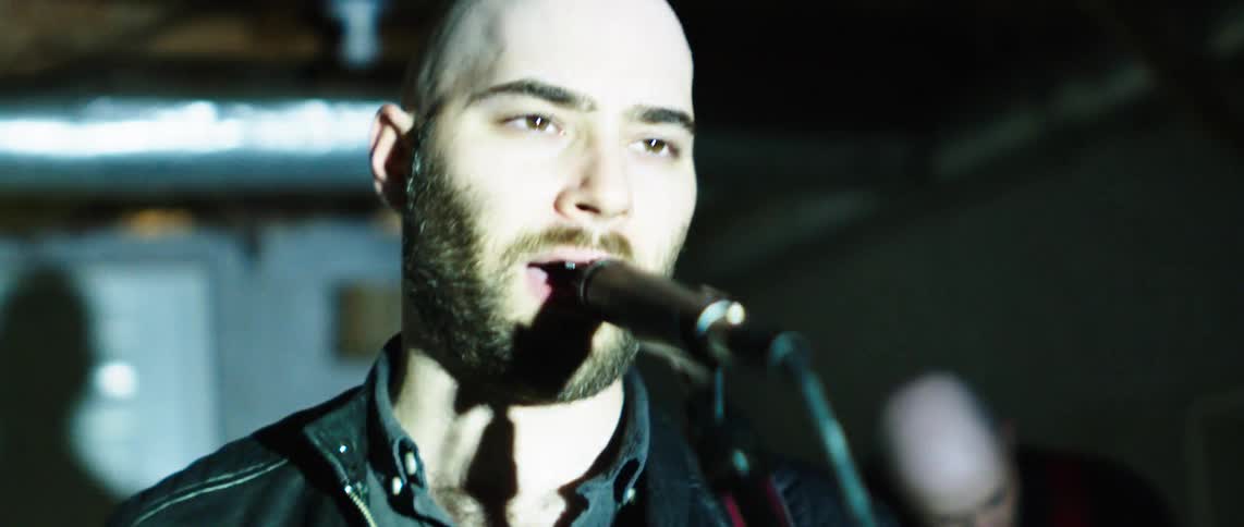 A bald man with a beard sings into a microphone while performing with his rock band on a stage with flashing lights