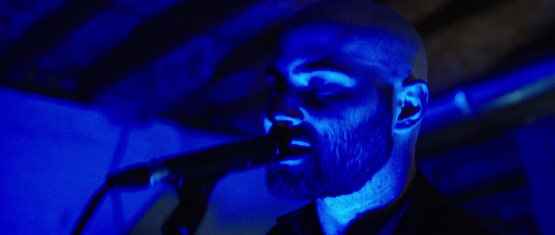 Two male guitarists play while in a room lit by colored lighting and one of the two musicians sings