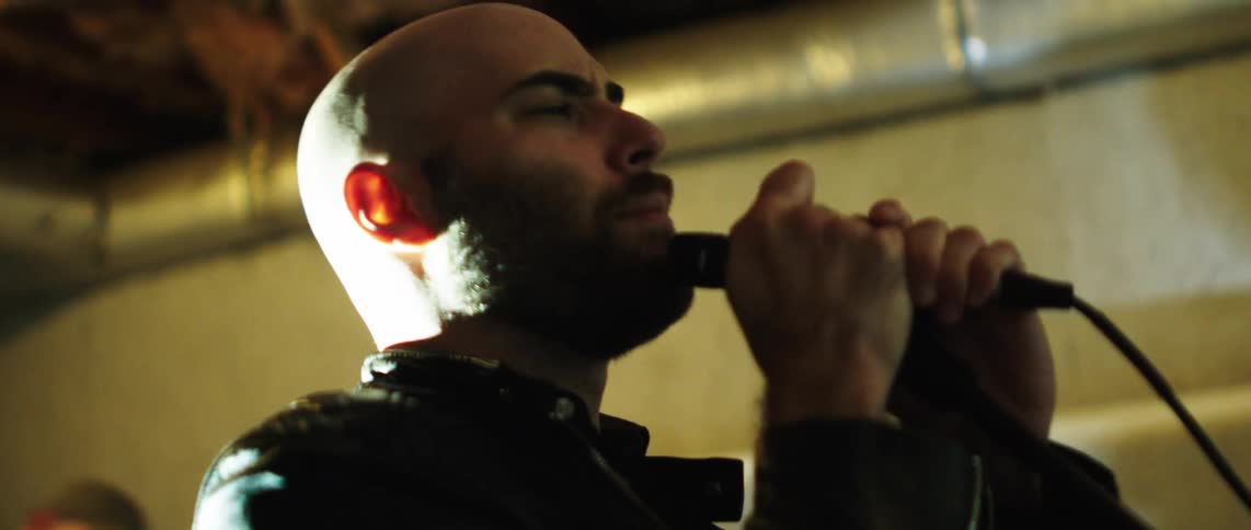 A bald man with a beard grips a microphone and stares intensely forward under dramatic pink lighting